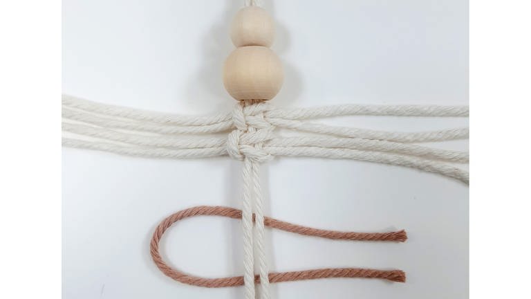 Nun das erste 20 cm Garn (beige) mittig falten und die Schlaufe nach links unter die beiden Fäden mit den Perlen schieben.  (Foto: Lisa Vöhringer)