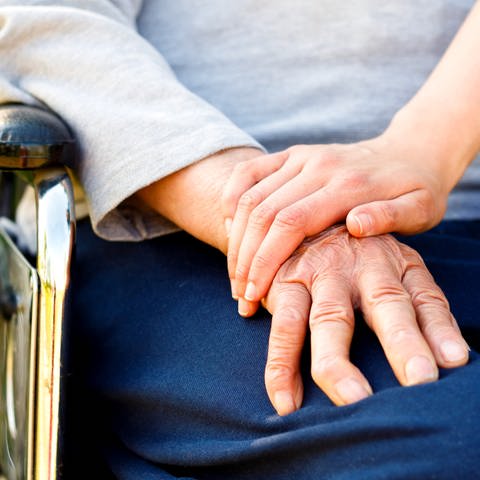 Hand eines älteren Menschen, der im Rollstuhl sitzt, auf dieser Hand eines jungen Menschen abgelegt (Foto: SWR)