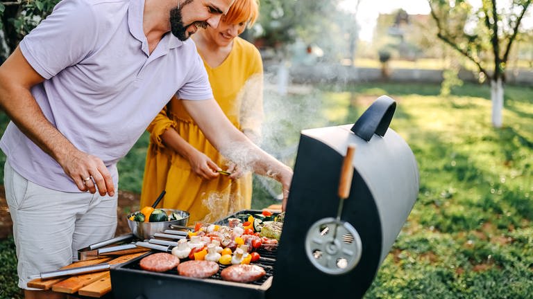 Mann und Frau stehen am vollbepackten Grill: Nachhaltige Alternativen zur Holzkohle (Foto: Getty Images, StefaNikolic)