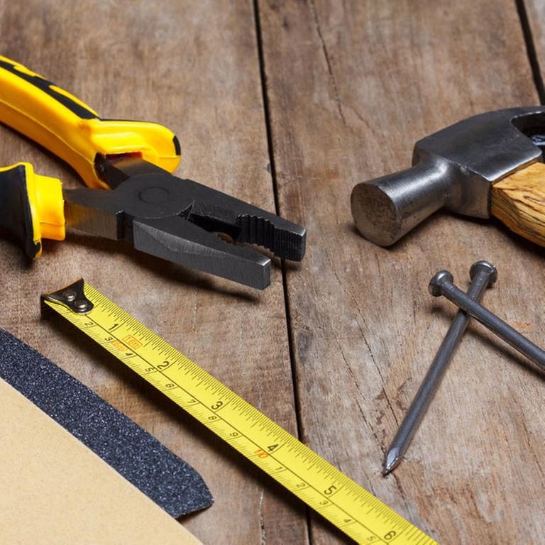Verschiedene Werkzeuge wie Zange, Hammer, Nägel, Metermaß und Schleifpapier liegen auf einem Holztisch.