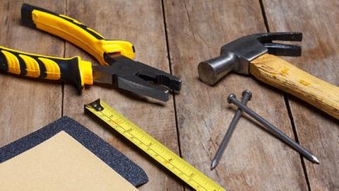 Verschiedene Werkzeuge wie Zange, Hammer, Nägel, Metermaß und Schleifpapier liegen auf einem Holztisch. (Foto: Colourbox, Foto: Colourbox.de -)