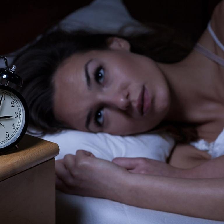 Frau liegt im Bett und schaut auf ihren Wecker der vier nach drei Uhr nachts zeigt.