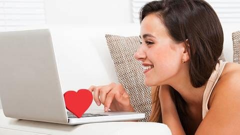 Lachende Frau liegt vor einem Laptop. Auf der Tastatur steht ein rotes Herz. (Foto: Getty Images, Thinkstock -)