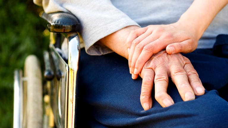 Hand eines älteren Menschen, der im Rollstuhl sitzt, auf dieser Hand eines jungen Menschen abgelegt