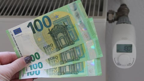IMAGOAnke Bingel (Foto: IMAGO, Energiepauschale: Eine Frau haelt drei 100-Euro-Geldscheine vor einen Heizkoerper)