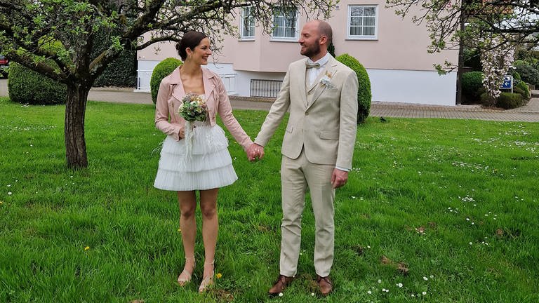Paar im Garten zeigt Brautkleid und Anzug für die Hochzeit (Foto: Brautsalon Lecher)