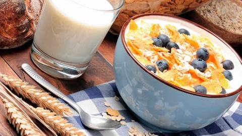 Ein Frühstückstisch mit einer Schale Müsli mit Obst, einem Glas Milch, und Brot.