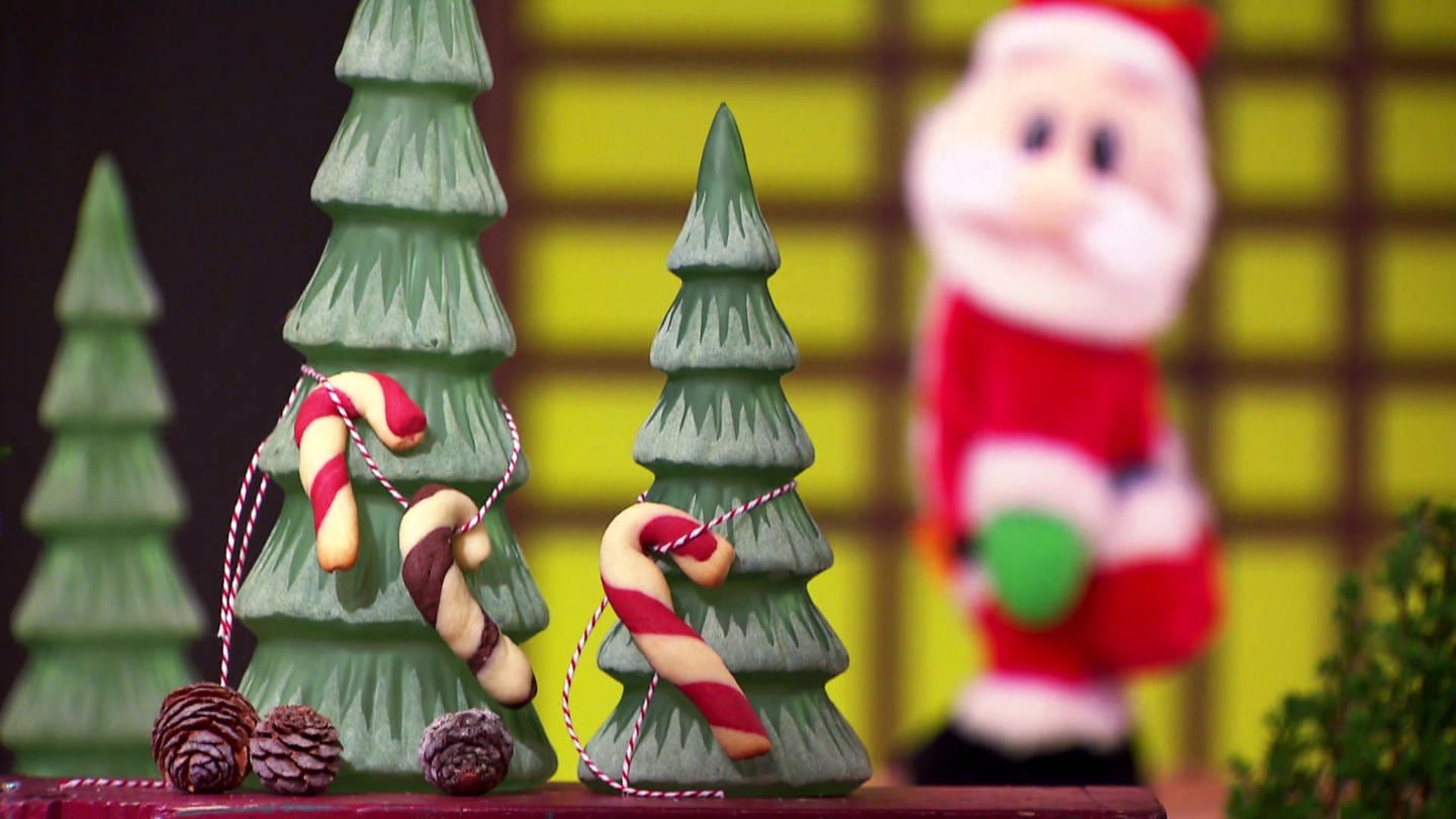 Drei Zuckerstangen-Cookies aufgehängt an zwei Deko-Tannenbäumen, im Hintergrund beleuchtete Weihnachtsmannfigur (Foto: SWR)