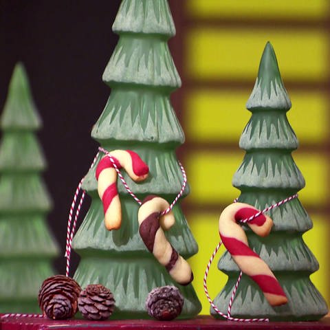 Drei Zuckerstangen-Cookies aufgehängt an zwei Deko-Tannenbäumen, im Hintergrund beleuchtete Weihnachtsmannfigur (Foto: SWR)