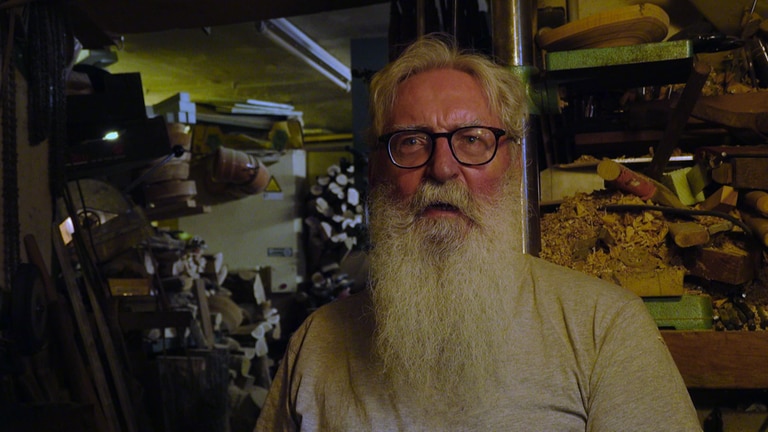 Ein älterer Mann mit langem Rauschebart steht in seiner Werkstatt voller Holz
