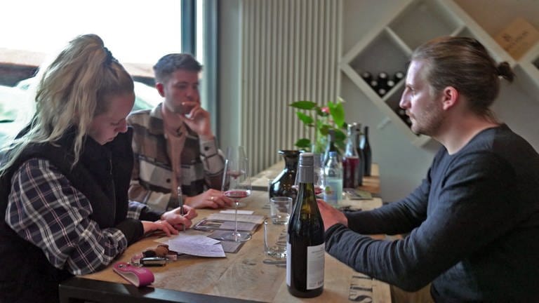 Annika und Manuel Braun sitzen bei einer Weinprobe mit einem Winzer am Tisch