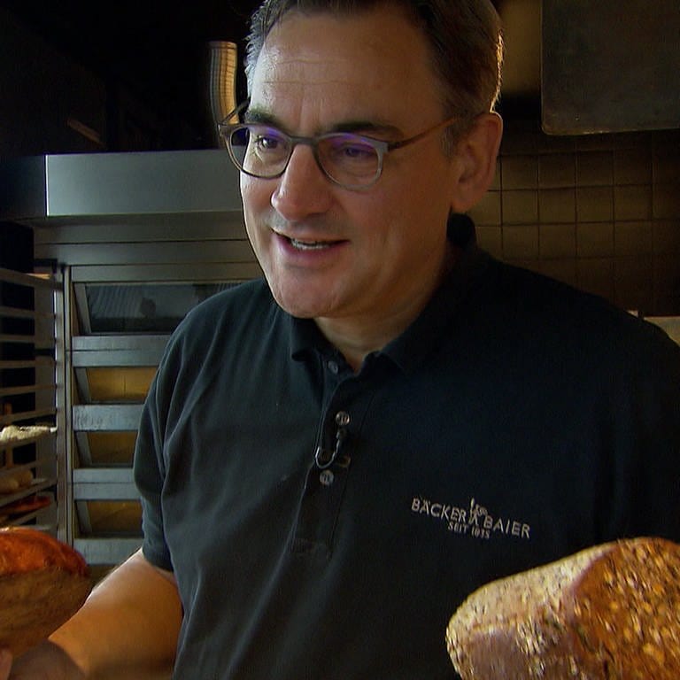 Bäcker Baier steht mit einem Brot in der Hand in seiner Bäckerei.