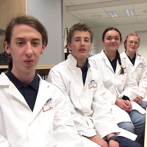 Vier Schüler in Laborkleidung sitzen nebeneinander an einem Tisch
