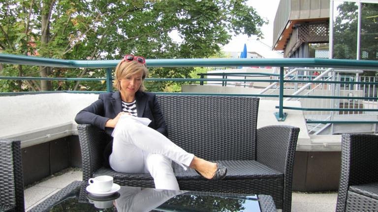 Moderatorin Annette Krause sitzt auf Korbsofa auf Terrasse, vor sich eine Kaffeetasse, und notiert etwas auf einem Schreibblock