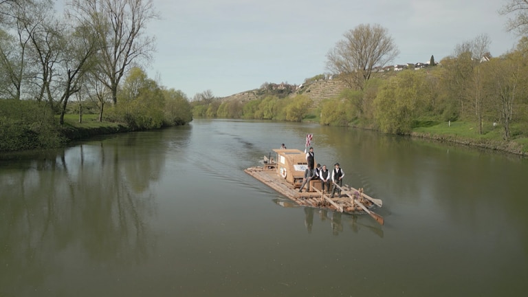 Auf einem hölzernen Floß fahren mehrere Männer den Neckar entlang.