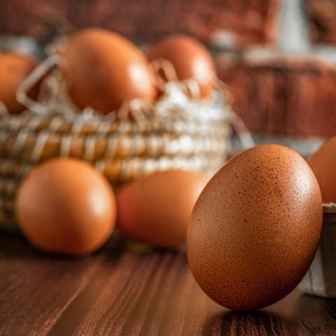 Im Vordergrund lehnt ein braunes Ei an einem vollen Eierkarton. Im Hintergrund sind weitere Eier in einem Strohkorb zu sehen. (Foto: Adobe Stock)
