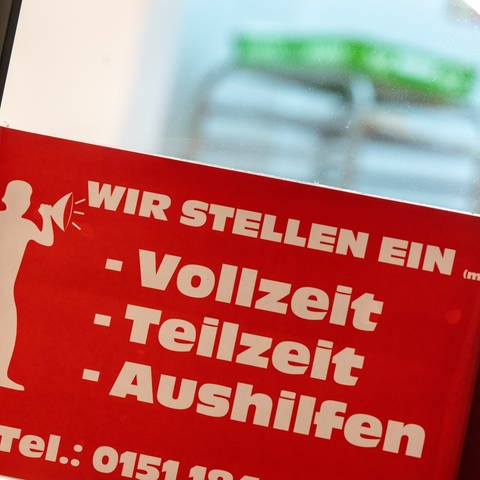 Ein rotes Schild mit der Aufschrift „Wir stellen ein - Vollzeit, Teilzeit, Aushilfen„ hängt am Eingang einer Bäckerei in der Innenstadt.