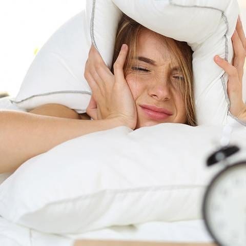 Eine Frau liegt im Bett und hält sich mit müdem Blick auf einen Wecker das Kopfkissen über die Ohren. (Foto: Getty Images, Thinkstock -)