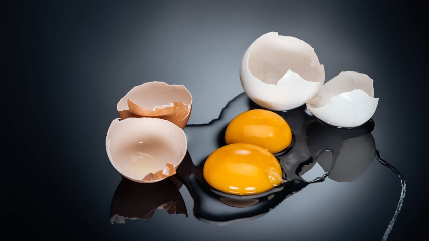 Zwei aufgeschlagene Eier liegen auf einem Tisch. Das Eiweiß ist rund um das Eigelb verteilt, diie Eierschalen liegen daneben.