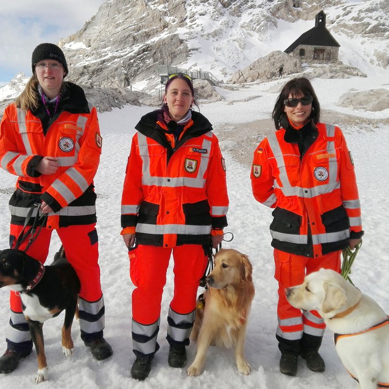 Drei Frauen von der Hundestaffel der Zugspitze stehen in orangenen Jacken mit Spürhunden vor einem Berg. (Foto: SWR)
