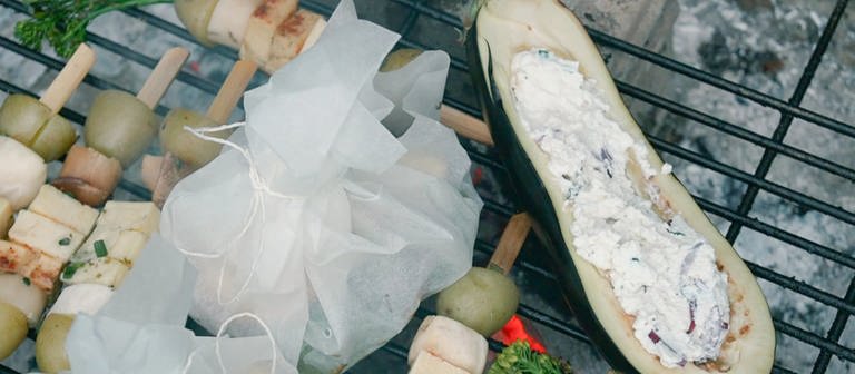 Eine gefüllte Aubergine, ein Gemüse-Käse-Spieß und gefüllte Backpapiertaschen liegen auf einem glühenden Grill. (Foto: SWR)