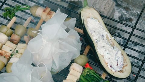 Eine gefüllte Aubergine, ein Gemüse-Käse-Spieß und gefüllte Backpapiertaschen liegen auf einem glühenden Grill. (Foto: SWR)