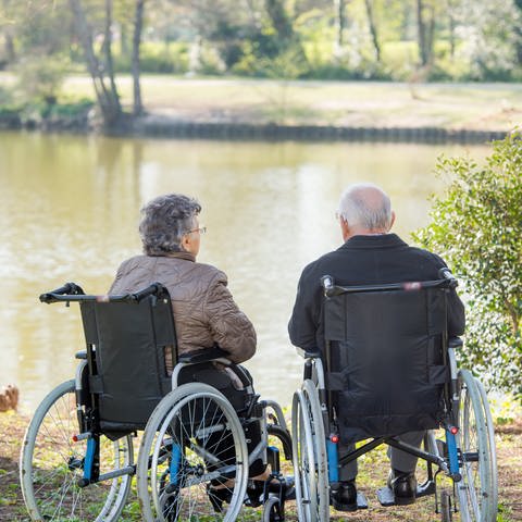 Ein älterer Mann und eine ältere Frau sitzen im Rollstuhl an einem See, der von einer grünen Wiese und Bäumen umgeben ist. (Foto: Colourbox, COLOURBOX25966188)