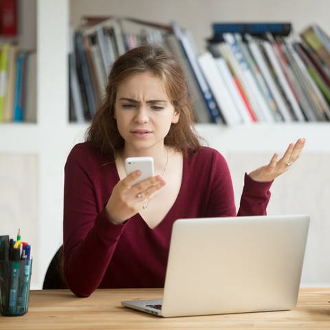 Junge Frau vor Laptop schaut verärgert auf ihr Handy (Foto: Colourbox, Colourbox/Aleksandr)