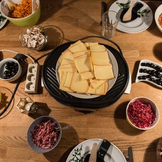 Raclettekäse und Schalen mit Zutaten stehen auf einem Tisch. (Foto: dpa Bildfunk, picture alliance / Patrick Seeger/dpa | Patrick Seeger)