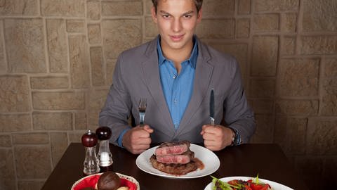 Junger Mann sitzt vor Teller mit drei Steaks. Männder sind oft mit Eisen überversorgt. Was kann man bei Eisenmangel tun? (Foto: Colourbox)
