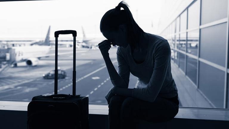 Eine Frau sitzt neben einem Koffer, verzweifelt mit dem Kopf auf eine Hand gestützt, vor einem Fenster am Flughafen. (Foto: Getty Images, getty images -)