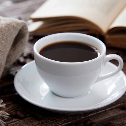 Eine Tasse Kaffe steht auf einem Tisch. Daneben liegen ein aufgeschlagenes Buch und eine Decke. (Foto: Colourbox)