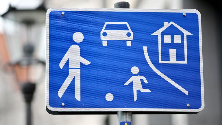 Auf eienm blauen Verkehrsschild sind  ein Mann, ein Kind, ein Ball, ein Auto, ein Haus und ein Straßenrand abgebildet. Es handelt sich um ein Schild, das eine verkehrsberuhigte Zone ankündigt, umgangssprachlich auch Spielstraße genannt. (Foto: picture-alliance / Reportdienste, Jan Woitas/ dpa)