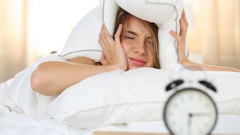 Eine Frau liegt im Bett und hält sich mit müdem Blick auf einen Wecker das Kopfkissen über die Ohren. (Foto: Getty Images, Thinkstock -)