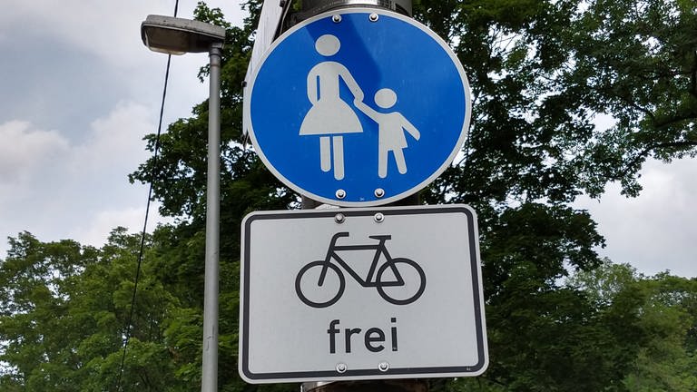 An einer Laterne hängt ein blaues Schild, das einen Fußgängerweg ausweist. Darunter befindet sich ein weißes Schild, das Fahrradfahrer frei bedeutet. (Foto: SWR, Thomas Oberfranz)