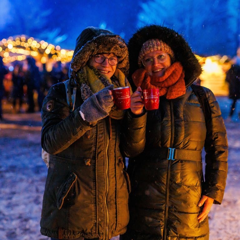 Zwei Frauen stehen mit zwei Tassen Glühwein in der Hand auf dem Weihnachtsmarkt.