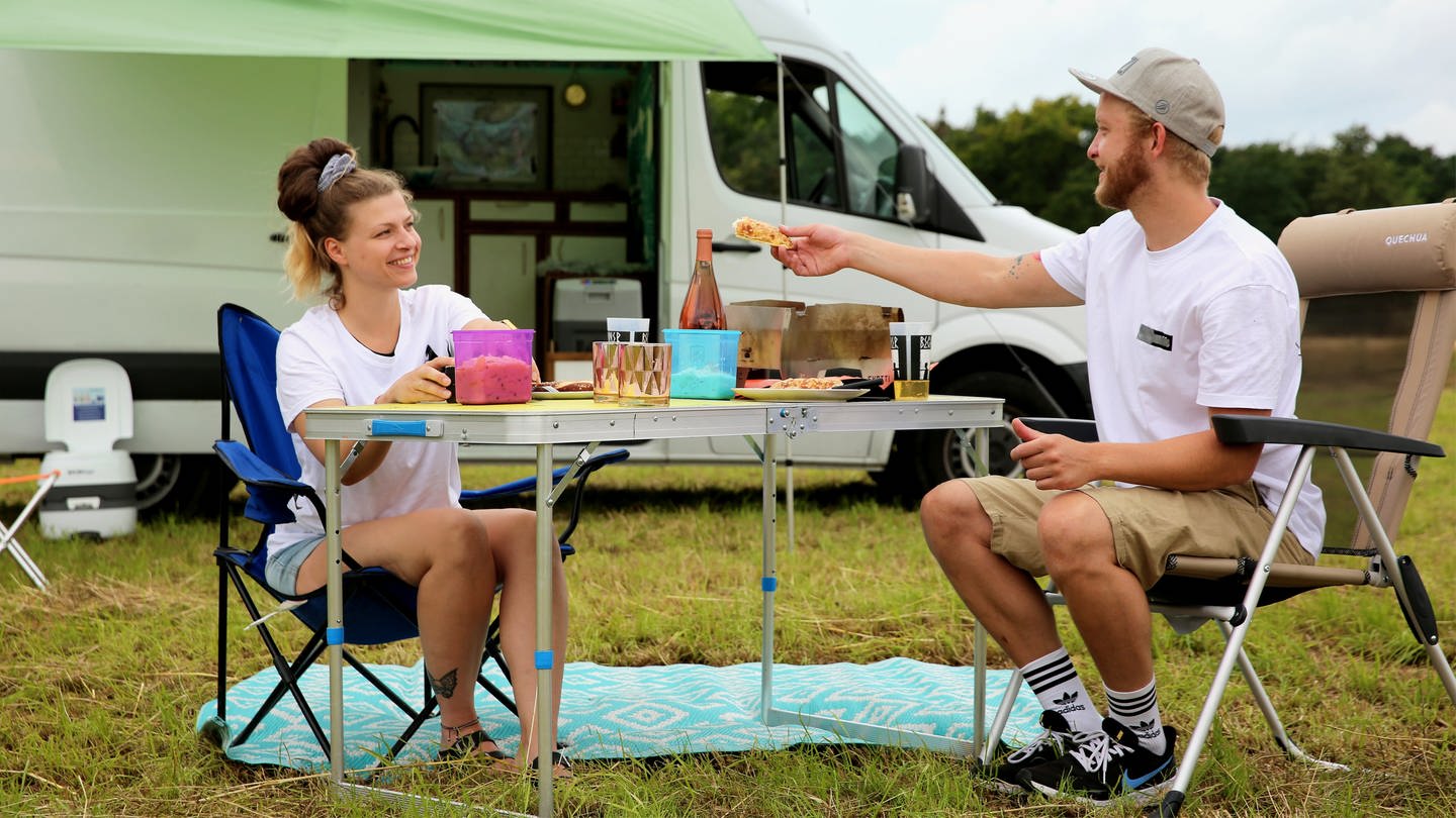 Von Campingbus bis Luxusvan: Zwei Personen sitzen vor einem Campingvan an einem gedeckten Campingtisch. (Foto: SWR)