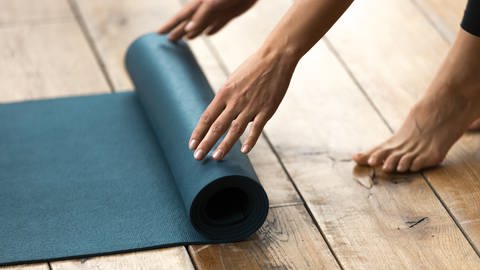 Eine Frau rollt eine Yogamatte auf dem Boden aus. Was sollte man beim Fitnessmatten-Kauf beachten?