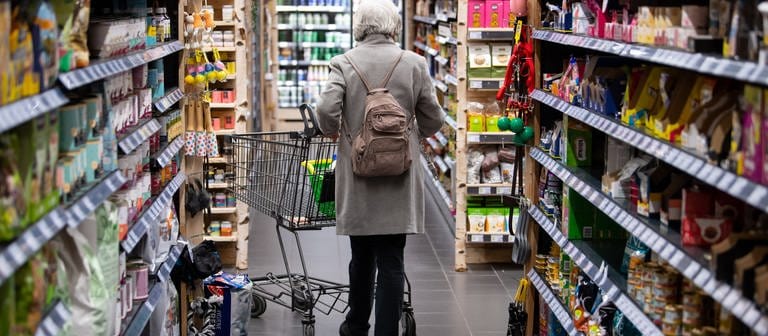 Ältere Frau von hinten gesehen schiebt einen Einkaufswagen an Supermarkt-Regalen vorbei. Preissteigerungen: Lebensmittel werden teurer (Foto: dpa Bildfunk, picture alliance/dpa | Sven Hoppe)