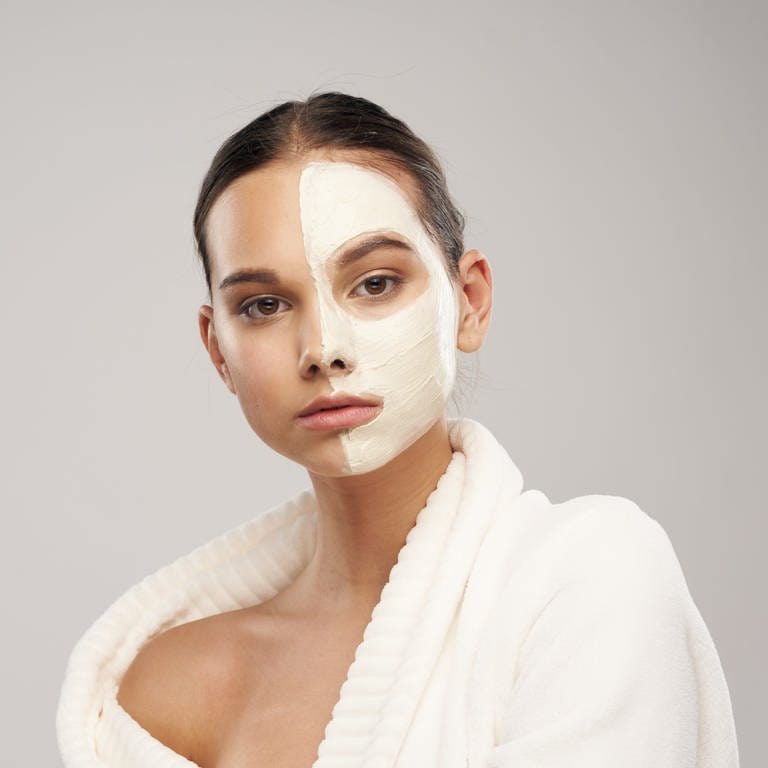 Junge Frau mit neutralem Blick und kosmetischer Creme-Gesichtsmaske, die nur auf einer Gesichtshälfte aufgetragen ist.  (Foto: Adobe Stock/SHOTPRIME STUDIO)