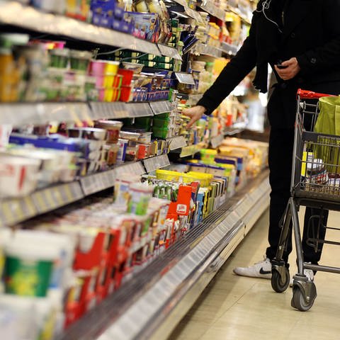 Person steht mit Einkaufswagen vor einem Supermarktregal und nimmt etwas heraus. (Foto: dpa Bildfunk, Picture Alliance)