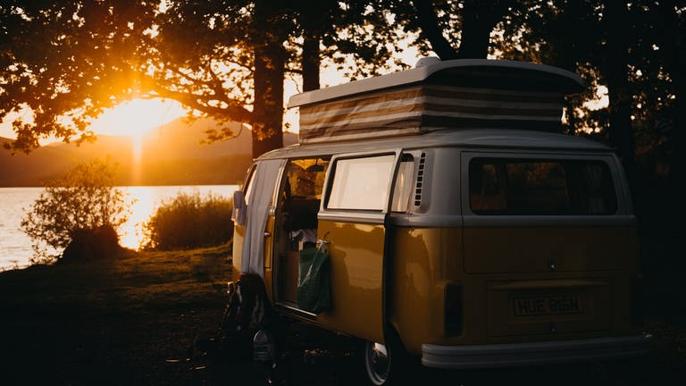 Alter Camperbus mit englischem Kennzeichen steht an einem See während die Sonne untergeht. (Foto: Unsplash/Kevin Schmid)