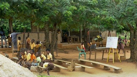 In einem ghanaischen Dorf sitzen Kinder auf Bänken im Freien. Im Rahmen seines "Farming Programs" baut Lindt & Sprüngli Schulen und Brunnen. (Foto: SWR)