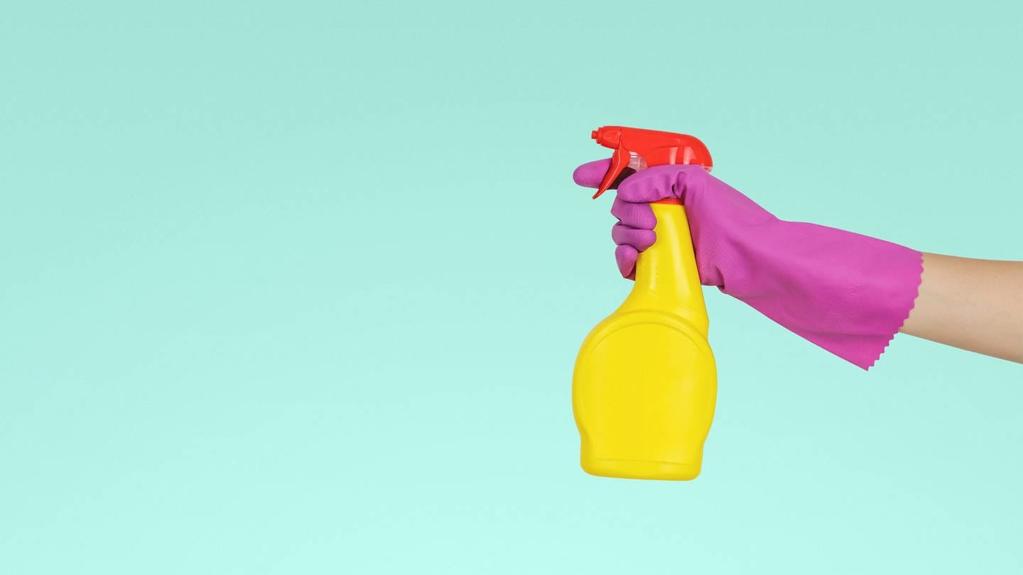 No-Name-Allzweckreiniger in grell gelber Sprühflasche wird von einer Hand in einem pinken Handschuh gehalten vor einer türkis-farbenden Wand.