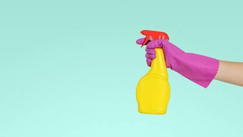 No-Name-Allzweckreiniger in grell gelber Sprühflasche wird von einer Hand in einem pinken Handschuh gehalten vor einer türkis-farbenden Wand. (Foto: Unsplash/JESHOOTS.COM )