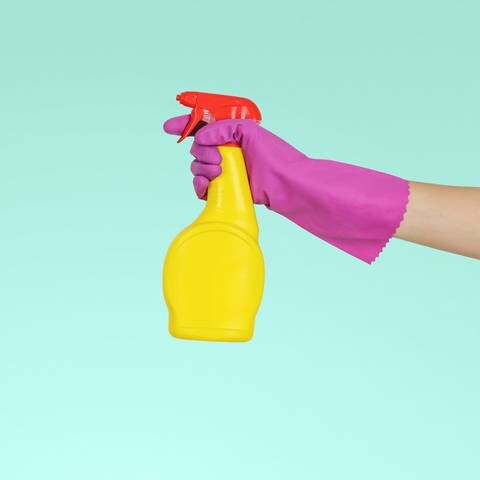 No-Name-Allzweckreiniger in grell gelber Sprühflasche wird von einer Hand in einem pinken Handschuh gehalten vor einer türkis-farbenden Wand. (Foto: Unsplash/JESHOOTS.COM )