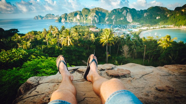 Post-Holiday-Blues: Urlauber fotostagrafit seine Beine mit Flips Flops an den Füßen auf einem Berg auf der thailändischen Insel Phi Phi. Die Aussicht kurz vor den Sonnenuntergang ist traumhaft schön.  (Foto: Unsplash(Evan Krause )