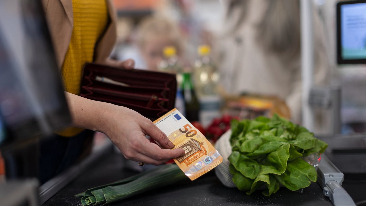 Person zahlt mit einem 50-Euro-Schein an der Kasser eines Supermarkts. Auf dem Kassenband liegt ein Salat und eine Lauchzwiebel, sowie weitere Produkte in der Unschräfe im Hintergrund. (Foto: Colourbox)