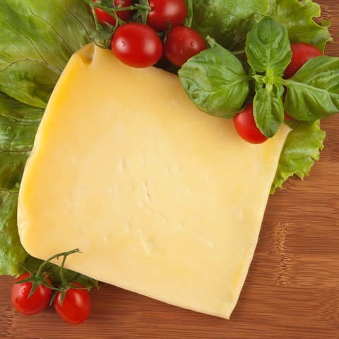 "Marke,Bio oder Discounter - Gouda im Produkttest" steht im Vordergrund auf einem semitranspartenen grünen Hintergrund. Im Hintergrund ist ein Stück Käse auf einem Holzbrett und mit Salatblättern und Tomaten garniert. In der rechten oberen Ecke ist die Bildquelle vermerkt. (Foto: Unsplash / Onder Ortel)