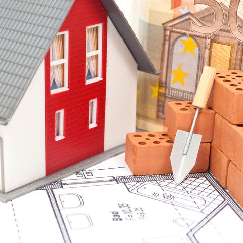 Symbolfoto: Auf einer Grundrisszeichnung steht ein Spielzeughaus, dahinter ein Geldschein, davor Ziegelsteine. Sind Bausparverträge als Geldanlage wieder attraktiver, seit die Zinsen steigen? (Foto: Colourbox)